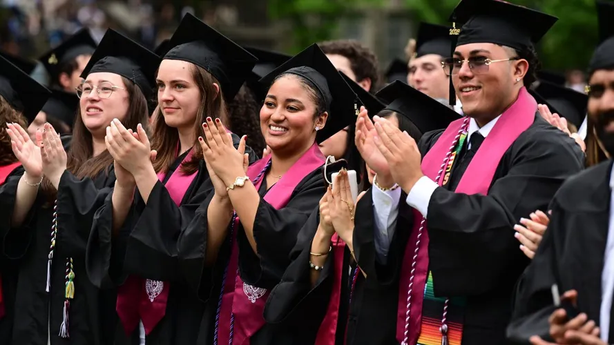 A group of students at graduation in caps and gowns stand and applaud during the ceremony.