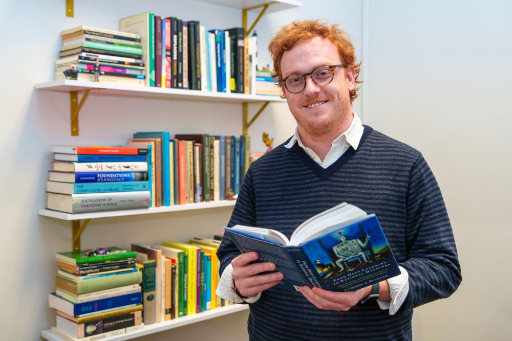 Sam McGrath stands in front of a book shelf with book in hand