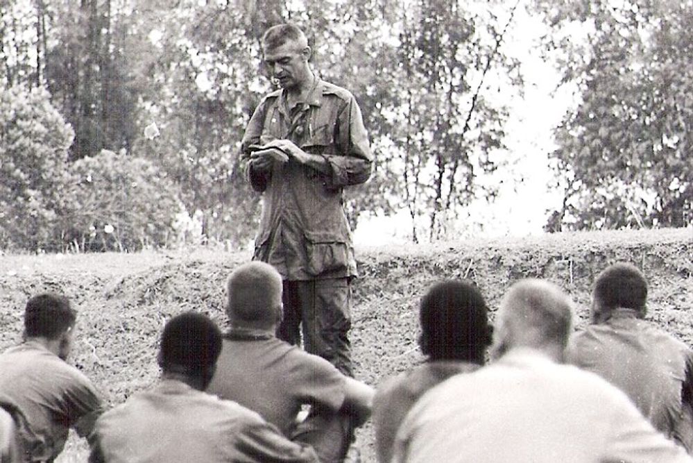 Fr. Capodanno standing in front of sitting Vietnam War troops with a small bible in his hand