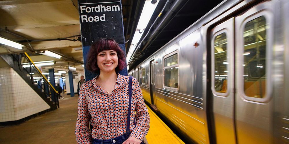 Female presenting student under Fordham Road sign in subway station. Subway to her left.