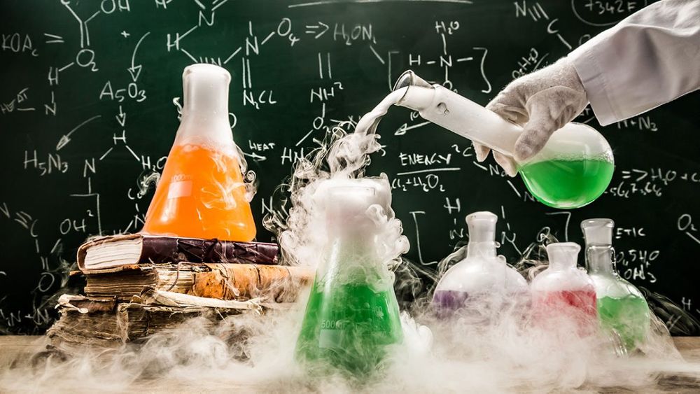 Several chemistry beakers filled with colorful liquids in front of a chalk board with chemistry formulas written on it