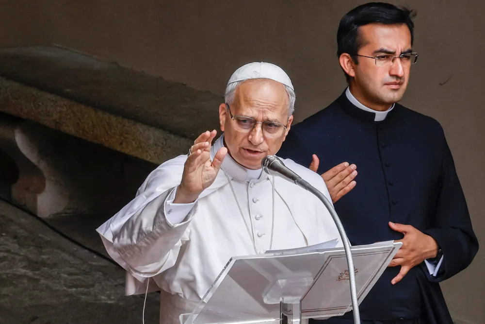 Pope Leo waves from the podium in white vestments as a priest stands behind him
