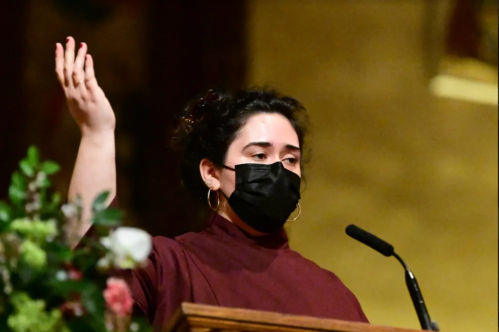 A student standing at an ambo with a mic in the University Church with her hand raised signaling for people to respond.