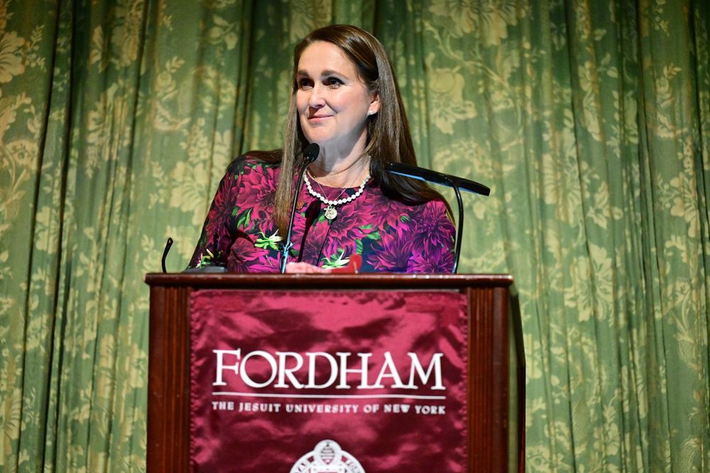 A female donor stands at a podium with a Fordham banner hanging while giving a speech at the Scholarship Donors and Recipients Reception