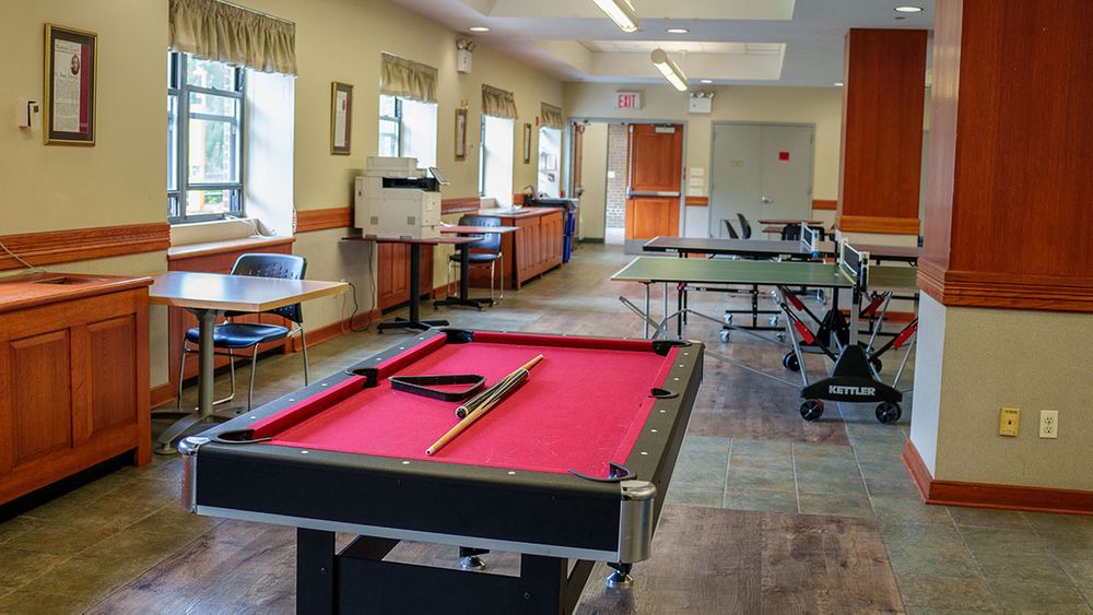 A red-felt pool table with cues and a cue stick rests in a well-lit recreational room featuring ping pong tables, desks, and windows.