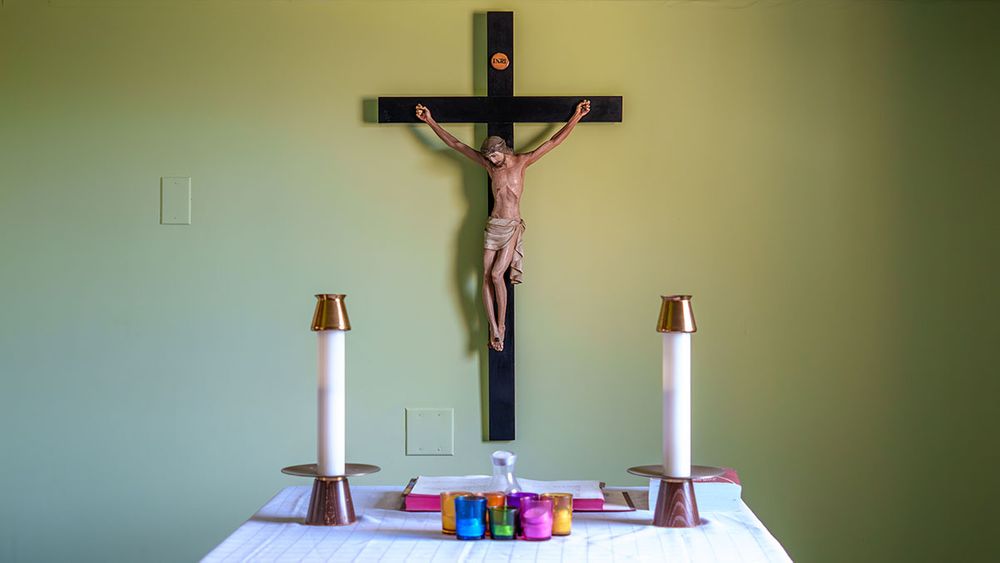 A crucifix mounted on a green wall above a table with candles and colorful containers.