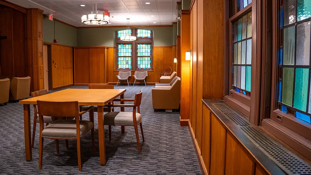 A warmly lit room with wooden paneling, stained glass windows, and a central table surrounded by chairs.