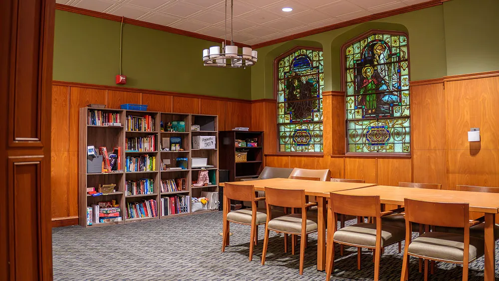A cozy room with wooden bookshelves, a long table with chairs, and two ornate stained glass windows set into green-walled space.