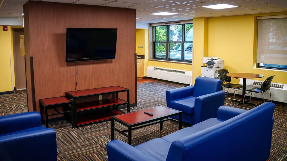 A modern lounge with blue upholstered seating, a mounted TV, and yellow walls, featuring a coffee table and a small dining area.