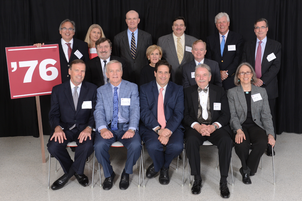 Alumni sit against black curtain holding red 76' sign