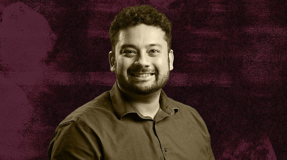A man with a beard wearing a dark shirt against a maroon textured background.
