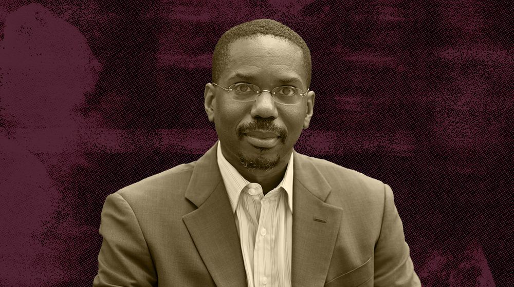 A man wearing glasses and a suit against a maroon textured background.
