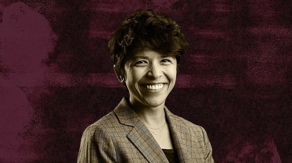 A woman with short hair wearing a patterned blazer against a maroon textured background.