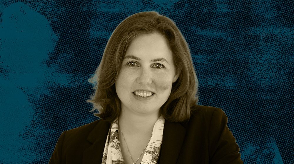 A woman with short hair wearing a patterned shirt and black blazer against a blue textured background.