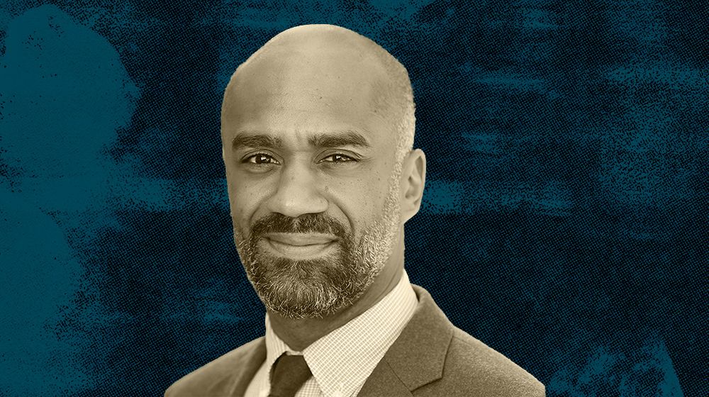 A man with a beard wearing a suit against a blue textured background.