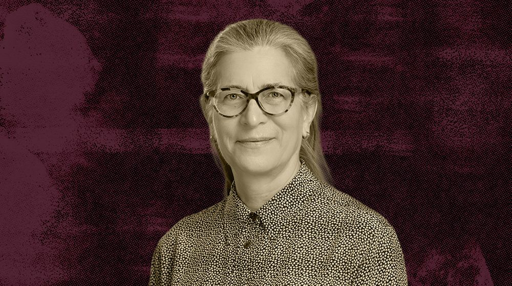 A woman with long hair wearing glasses and a patterned shirt against a maroon textured background.