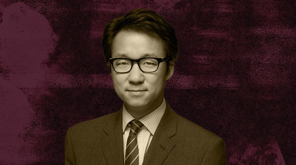 A man wearing glasses and a suit against a maroon textured background.