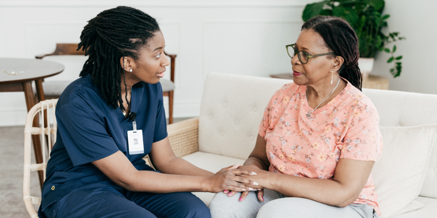 A healthcare worker in blue scrubs gently holding hands with an older woman sitting on a couch while they have a supportive conversation.