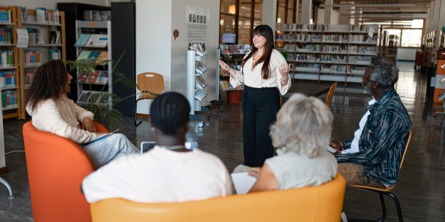 A woman stands and presents to a diverse group of people sitting in a circle of colorful chairs in a library.