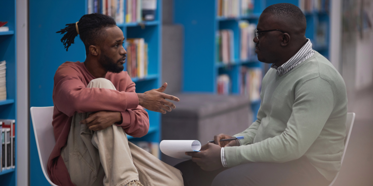 A school social worker holding a notepad and listening attentively to a young student speaking in a library.