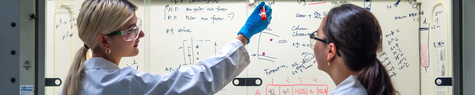 Two female students in labs coats writing on a white board