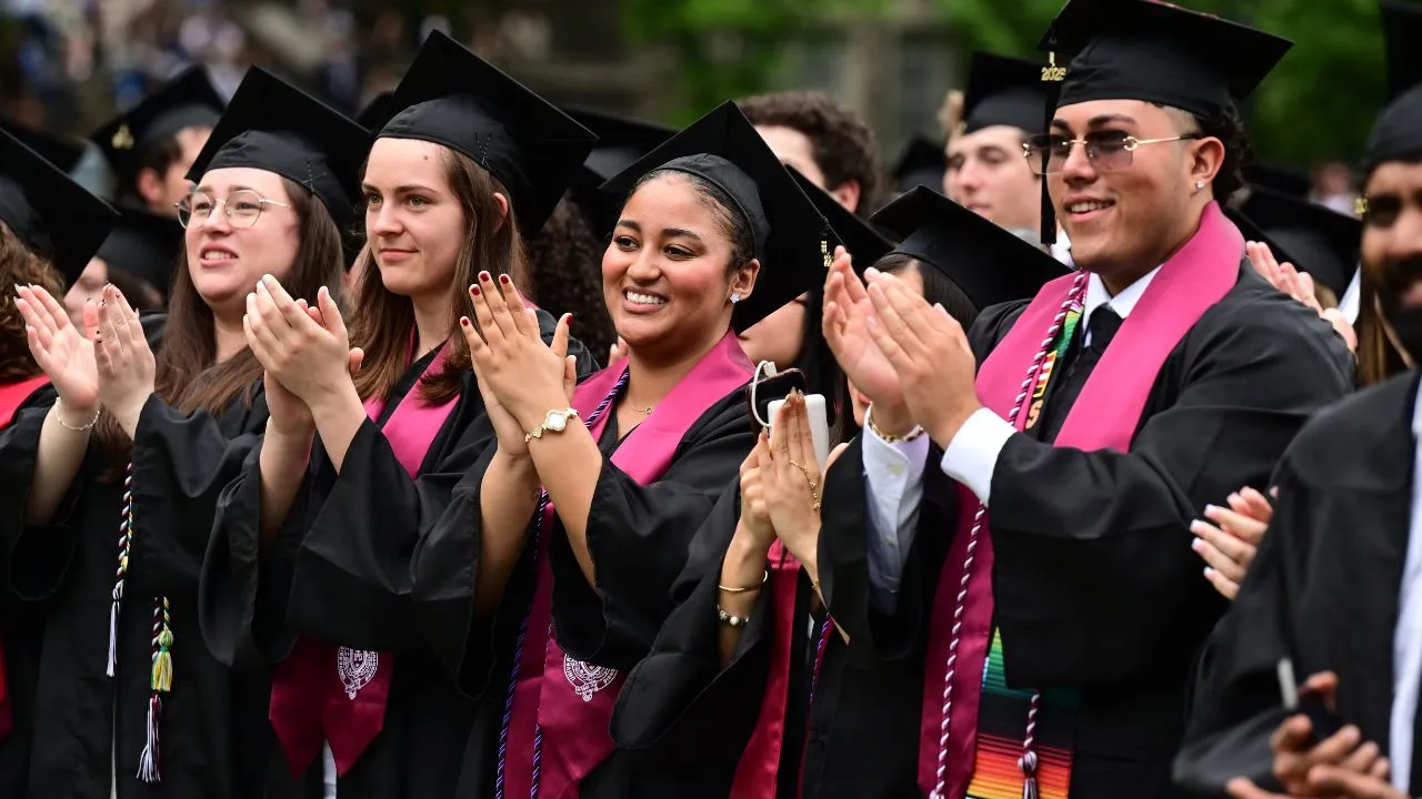 A group of students at graduation in caps and gowns stand and applaud during the ceremony.
