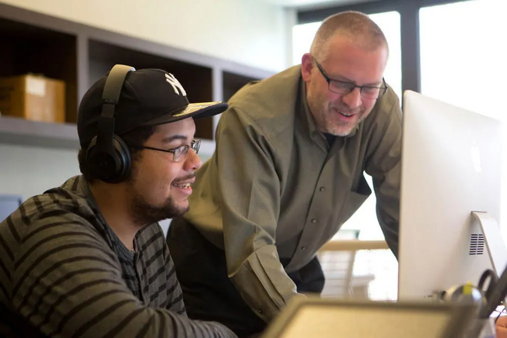 A male faculty member looks at a computer monitor with a student wearing a Yankees hat and headphones