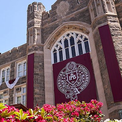 Keating hall unclose shot, Fordham seal hanging from Keating with red flowers in forefront.