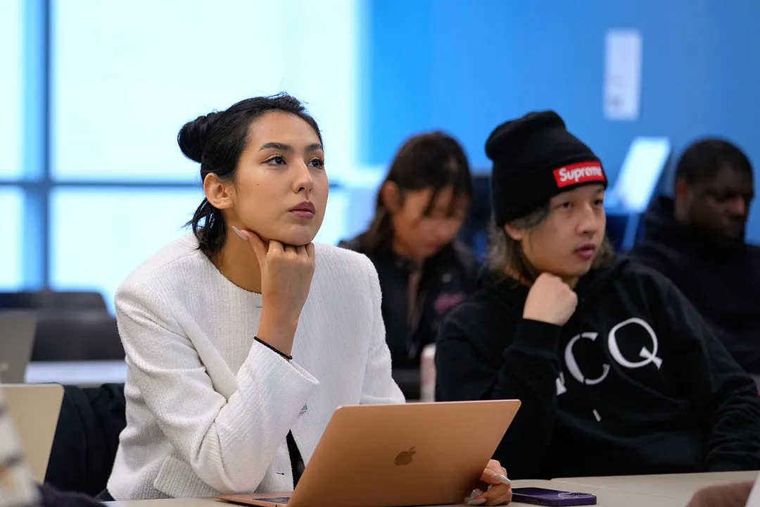 A female graduate student sits at a desk with a laptop in a classroom with other students behind her
