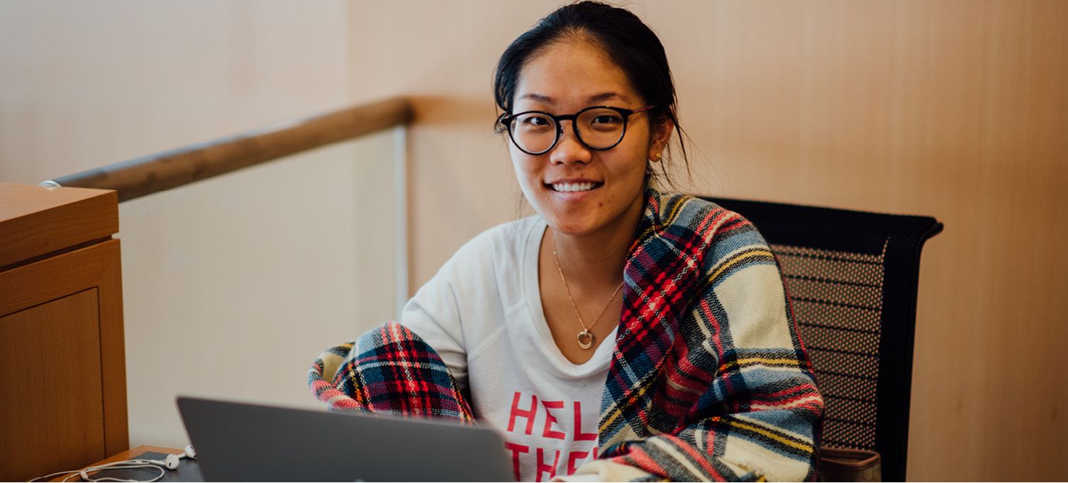 Female Student with Glasses Smiling at Camera while Using a Laptop.