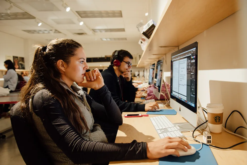 A female student concentrates on a computer screen in a lab setting with other students on computers