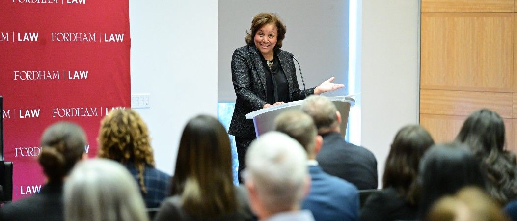 A woman wearing a suit standing at a podium speaking to a seated audience.