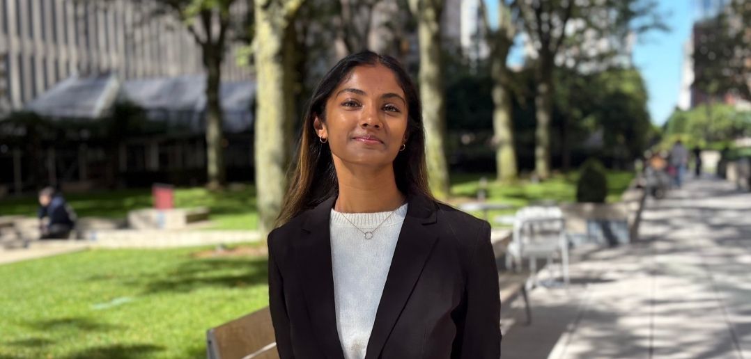 A woman wearing a white shirt and black blazer standing in front of a bench and trees.