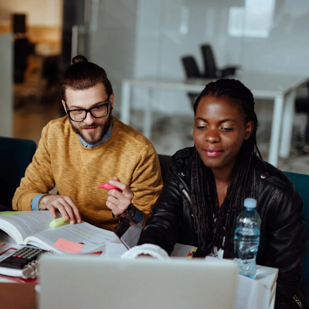 A man and a woman sitting at a desk with books, sticky pads, and a laptop.